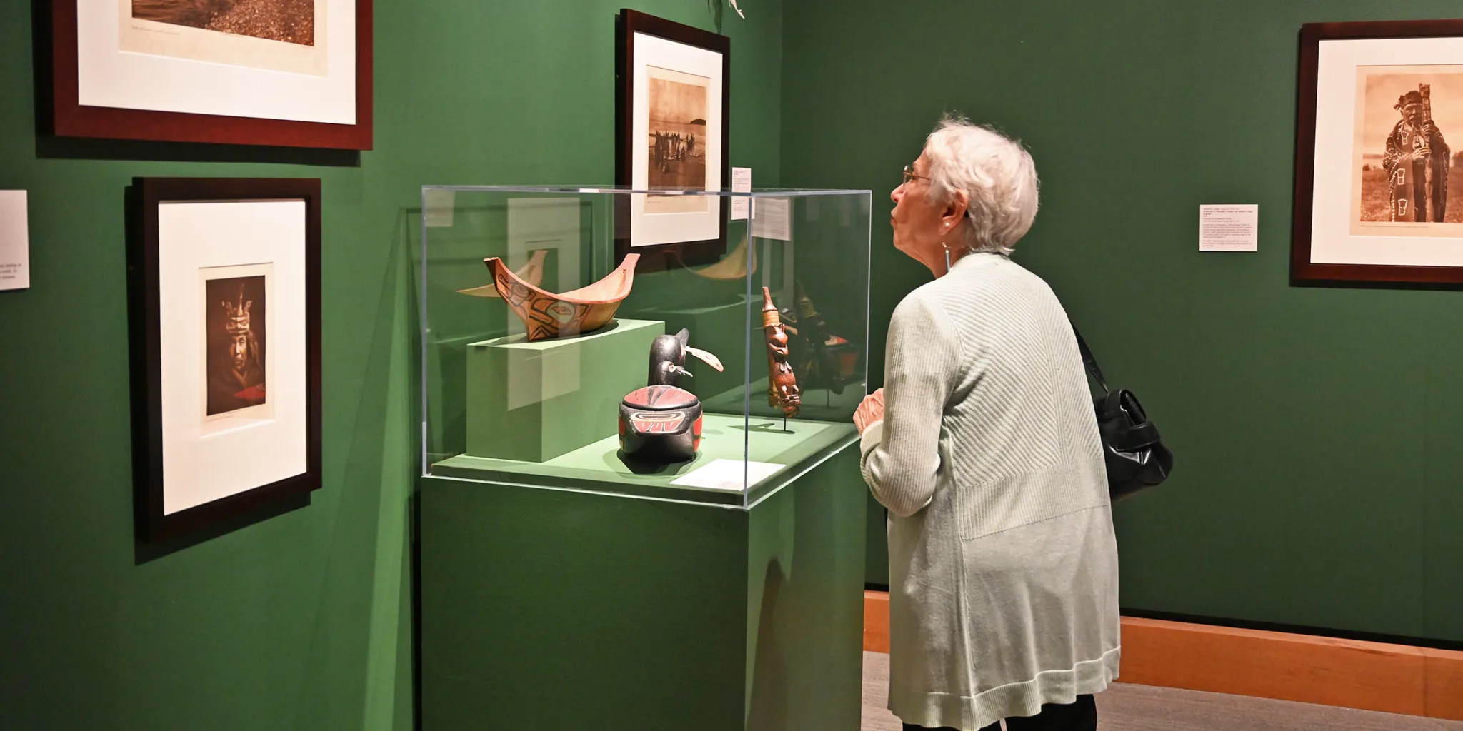 An older woman observes artifacts and framed photographs displayed in a museum exhibition with dark green walls, exploring the stories behind current exhibitions.