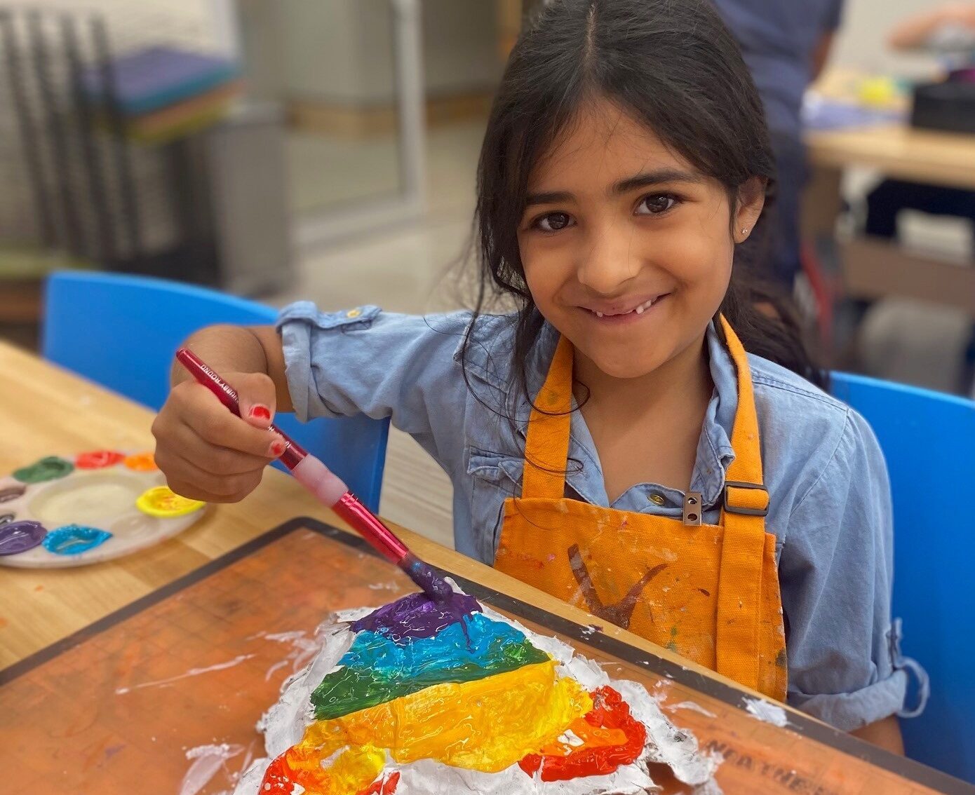 A young girl wearing an orange apron is painting a rainbow on a piece of paper at a table, smiling at the camera as she learns about financial literacy through creative activities.