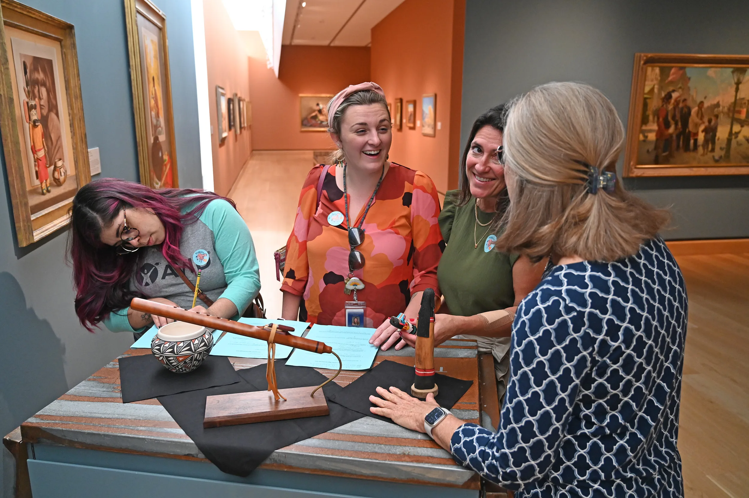 Four educators stand around a display table with various objects in an art gallery, smiling and talking together. Paintings hang on the walls in the background.