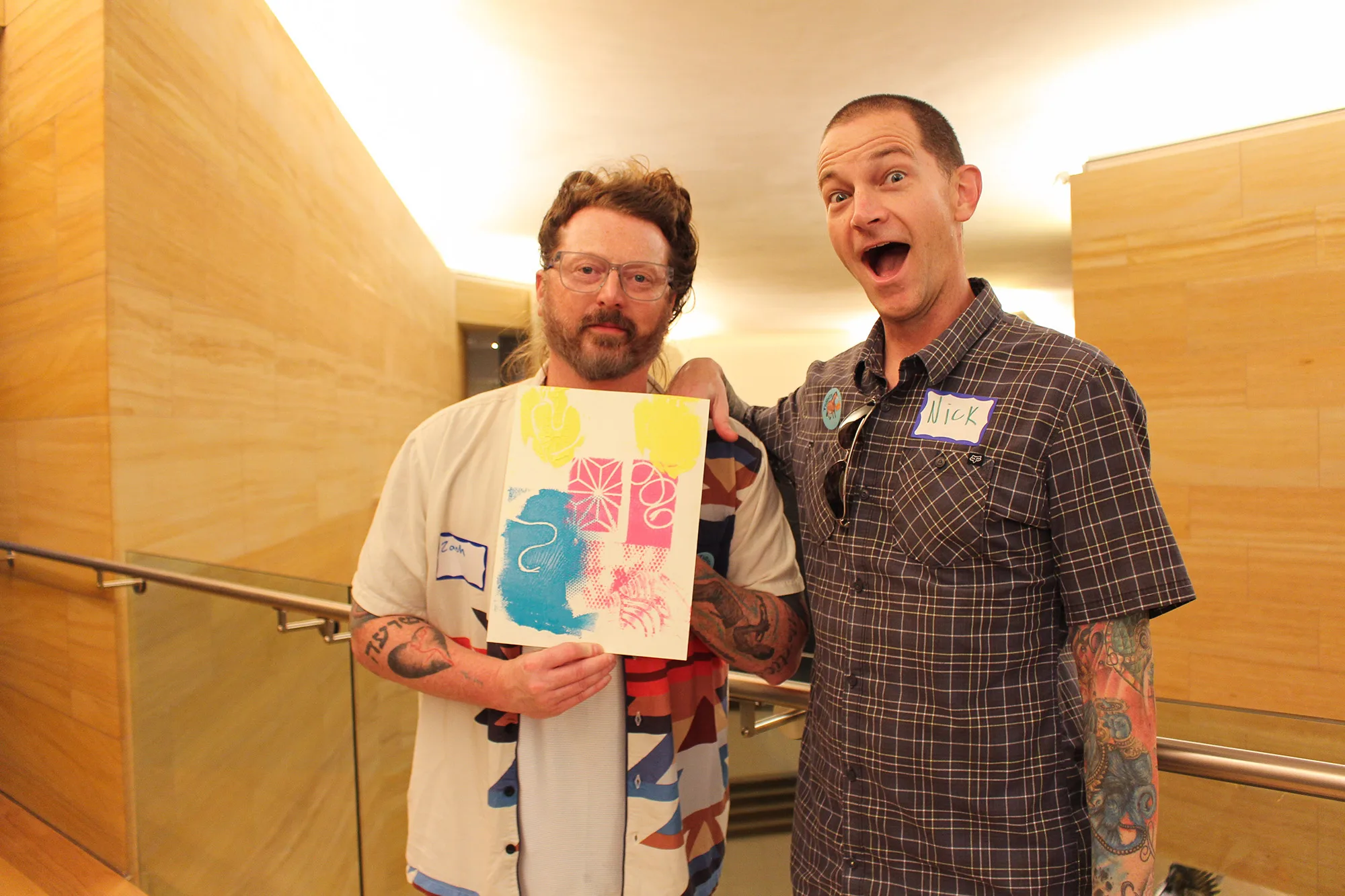 Two educators stand indoors, one holding a colorful abstract art piece. Both wear name tags and casual shirts, with a beige wall in the background.