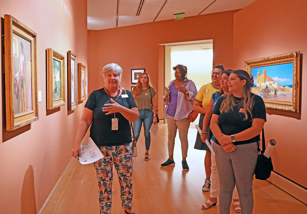 A museum guide, much like educators, leads a group of visitors through an art gallery, explaining the paintings displayed on the walls.