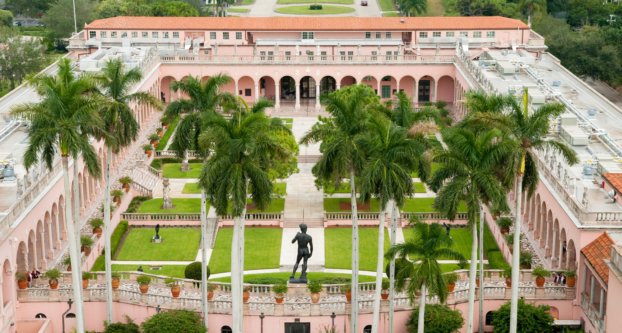 Aerial view of a pink historic building with arched walkways, a central courtyard, palm trees, and a statue in the garden—an exclusive destination where member benefits enhance your visit.