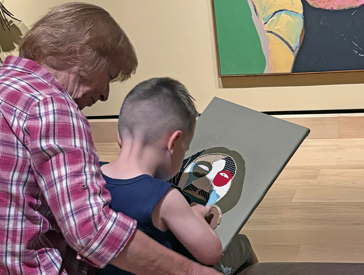 An adult and a child sit together as the child paints a colorful abstract face on a canvas in an art gallery, sparking conversations about creativity and financial literacy through the art’s depiction of money value.