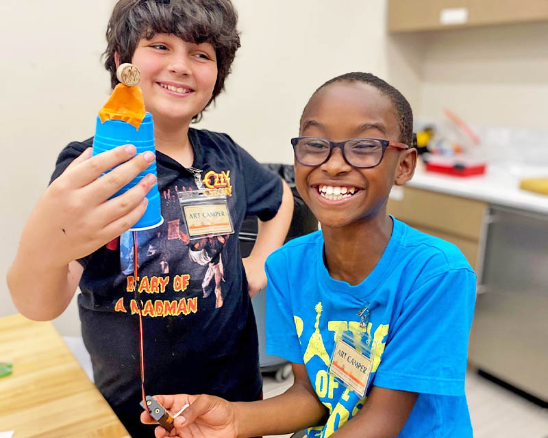 Two children smiling, one holding a homemade device with a coin attached by string, in a classroom or activity room setting.