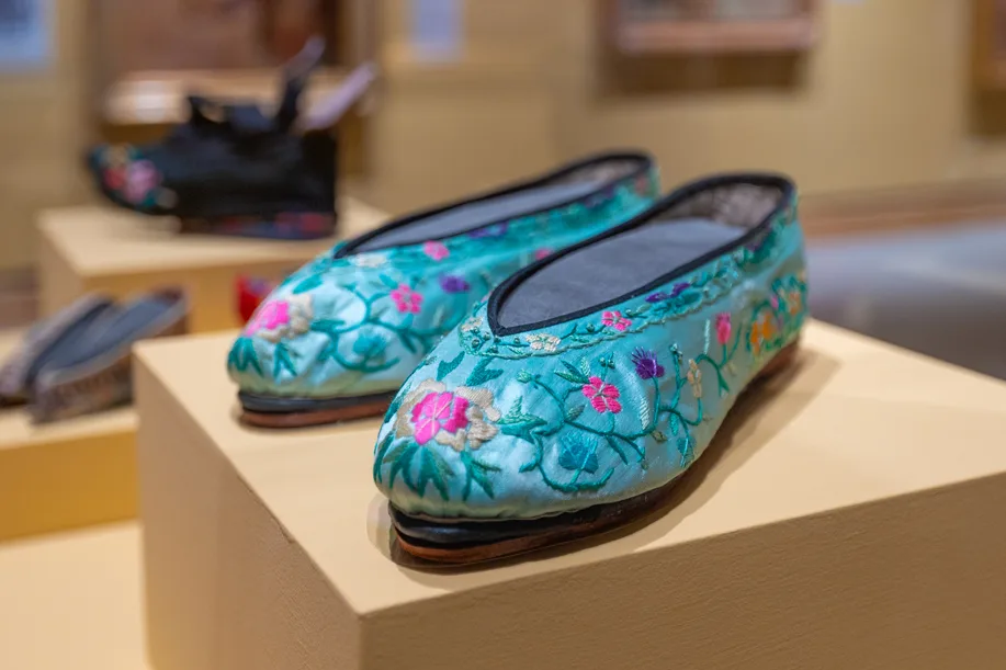 A pair of traditional Chinese embroidered silk shoes with floral patterns, reminiscent of FarEast 09, are displayed on a pedestal in a museum.