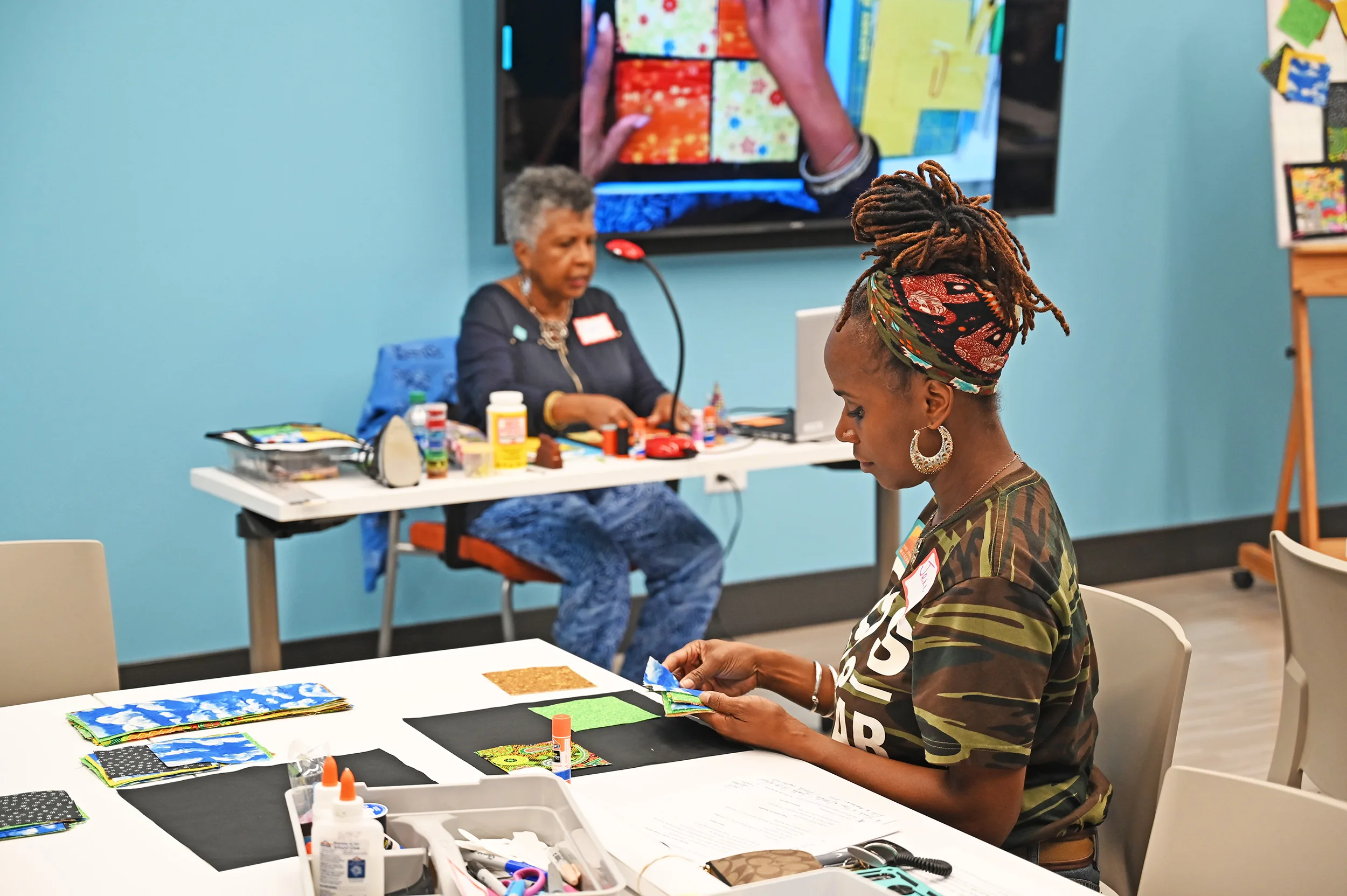 Two women participate in an arts and crafts activity as part of our Adult Programs; one works at a table with paper and glue, while another sits at a desk in the background.