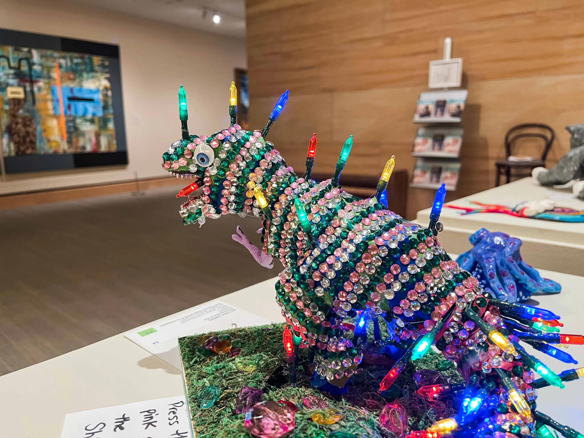 A small sculpture of a T-Rex bedazzled with rhinestones and string lights on a podium.